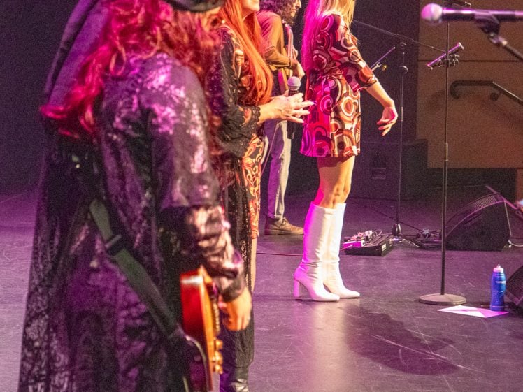 Image of Four musicians on stage, three women in colourful retro outfits and one man in the background. The women are at microphones, with one playing guitar. Stage lights highlight their vibrant costumes.