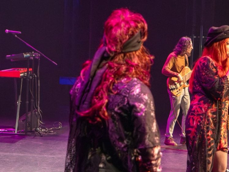 Image of A band performs on stage with a keyboard player on the left, a guitarist in the background, and two female singers in the foreground, all under purple and pink stage lighting.