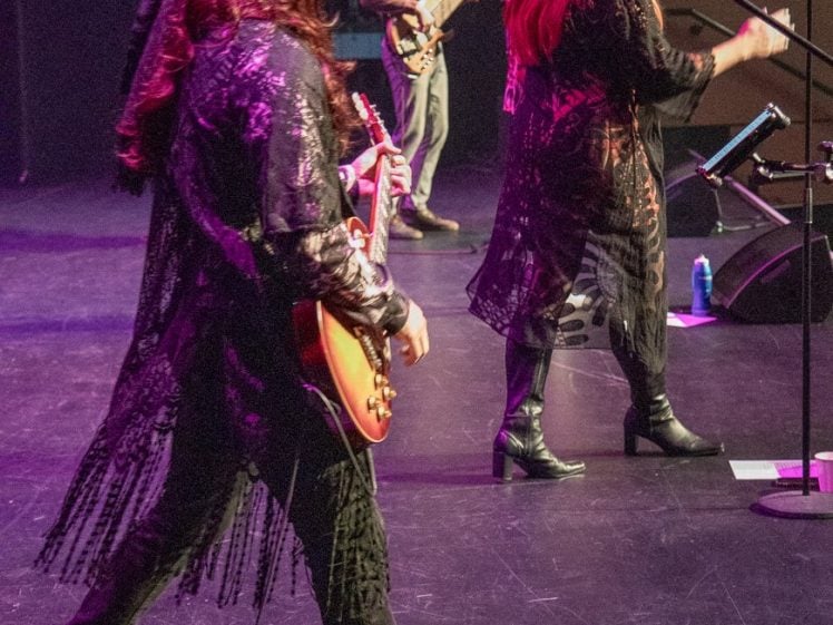 Image of Musicians in retro outfits perform on stage with guitars and microphones, colourful lighting shining on them. The focus is on a guitarist and a vocalist in fringe and patterned clothing.