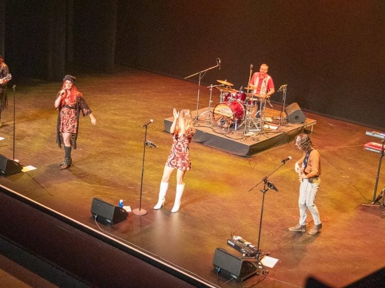 Image of A six-member band performs on a brightly lit stage. Two singers are at the front, with one clapping. The other members play electric guitar, bass, drums, and keyboard. Musical equipment is scattered around the stage.
