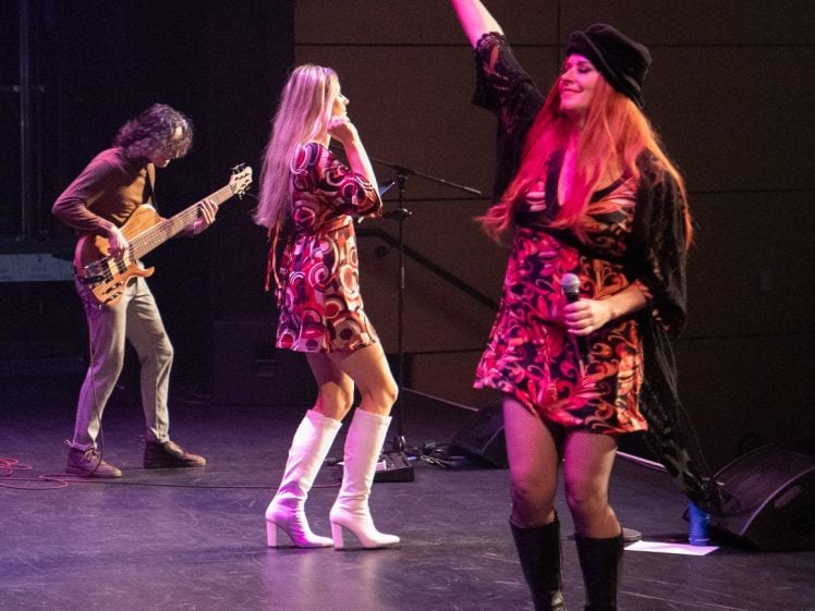 Image of Three musicians perform on stage; a guitarist in the background, a singer with long blonde hair and white boots in the centre, and a singer in front with long red hair raising one arm, all wearing retro-inspired outfits.