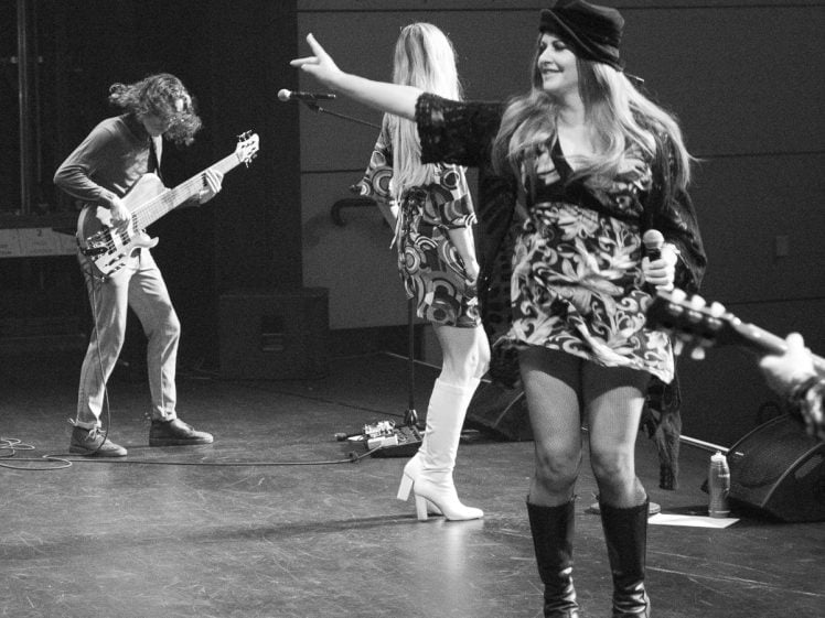 Image of A woman in a dress and boots sings and points while holding a microphone on stage with two musicians, one playing guitar and another in white boots, during a live performance. The image is in black and white.
