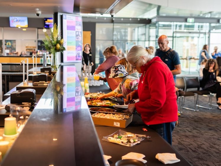 Image of People helping themselves to food from a buffet table in a modern indoor setting, with others seated and socialising in the background. The atmosphere appears casual and welcoming.