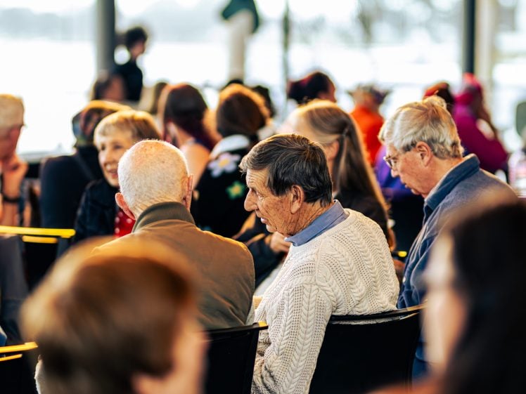 Image of A group of older adults sit closely together in a bright, indoor setting, engaged in conversation. The background is softly blurred, suggesting a social or community event.