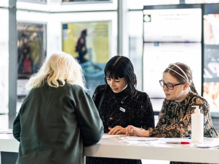 Image of Three women are standing at a counter, two facing forwards and one with her back to the camera. They appear to be engaged in conversation or paperwork, with posters and screens visible in the background.
