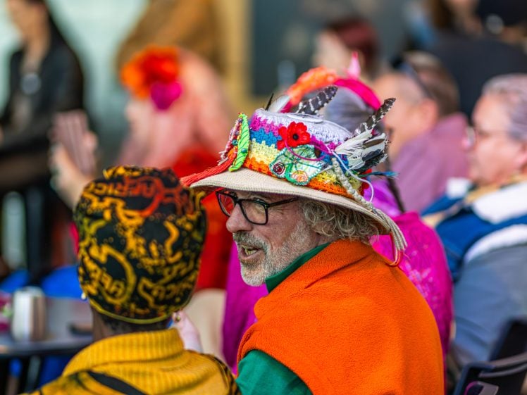 Image of A man wearing glasses, an orange cape, and a colourful hat decorated with flowers and feathers sits among a group of people, some with vibrant clothing and hats, at what appears to be a lively public event.