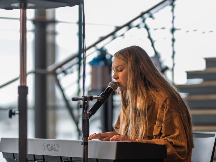 Image of A woman with long blonde hair plays a Yamaha keyboard and sings into a microphone. She is indoors near large windows and staircase bannisters, focused on her music.