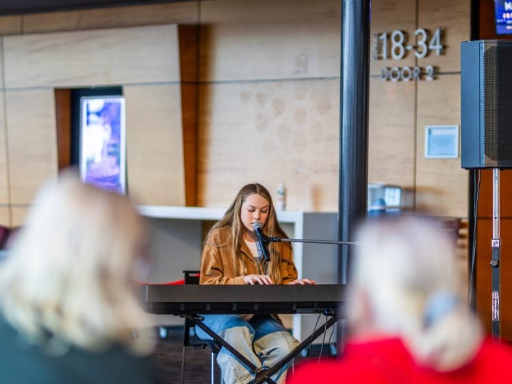 Image of A young woman plays a keyboard and sings into a microphone in an indoor venue, with two blurred people seated in the foreground watching her performance.