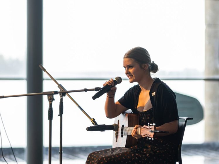 Image of A young woman sits on a chair indoors, holding an acoustic guitar and speaking or singing into a microphone, with a second microphone stand nearby and bright natural light in the background.