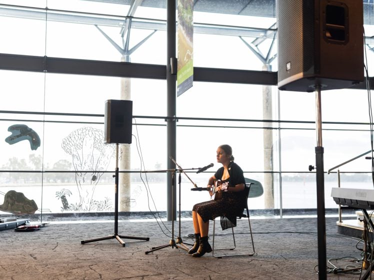 Image of A woman sits on a chair playing guitar and singing into a microphone at an indoor venue with large windows, loudspeakers, and natural light streaming in.