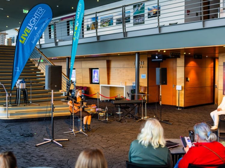 Image of A musician performs on stage with a guitar in a modern indoor venue, watched by an audience. Banners with Live Lighter branding are displayed beside the performer. Stairs and a railing are in the background.
