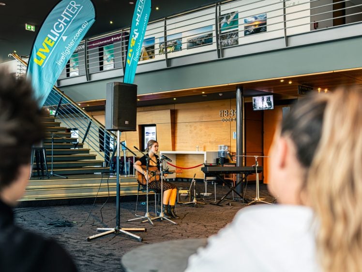 Image of A woman sits on a chair playing guitar and singing into a microphone in a modern indoor venue, with two people watching in the foreground. Banners reading LIVELIGHTER are displayed nearby.