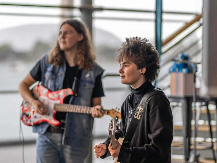 Image of Two young people play electric guitars indoors. The person in the foreground has spiky hair and wears a dark jacket, while the person in the background has long hair, a denim waistcoat, and is slightly out of focus.