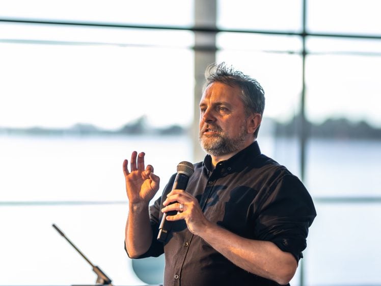 Image of A man with a beard and wearing a dark shirt speaks into a microphone, gesturing with his hand, whilst standing in front of large windows with a blurred outdoor view in the background.