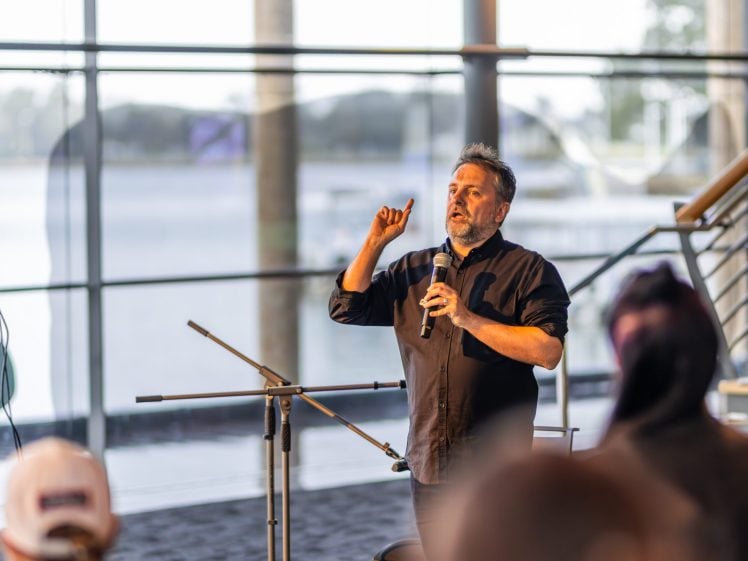 Image of A man holding a microphone gestures whilst speaking to an audience in a modern indoor space with large windows, overlooking a waterfront.