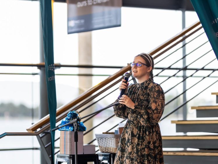 Image of A woman wearing glasses and a patterned dress speaks into a microphone on an indoor stage near a staircase, with a gift hamper and wrapped box on a nearby table.