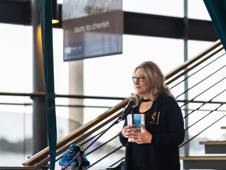 Image of A woman with long blonde hair and glasses stands by a staircase, speaking into a microphone and holding a book. She is wearing a black jacket and is indoors near large windows.