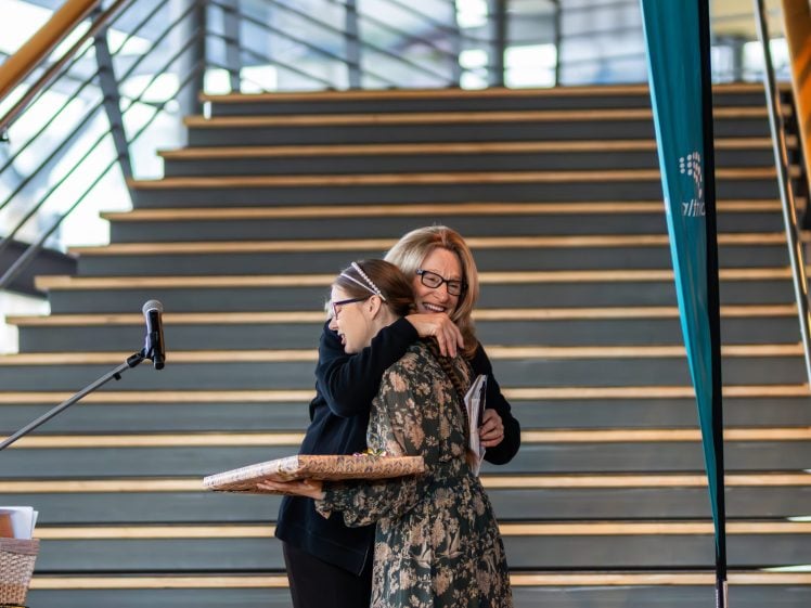 Image of Two women embrace and smile on a stage in front of wide steps. One woman holds a large wrapped present, and a microphone stands nearby, suggesting an award or celebration event.