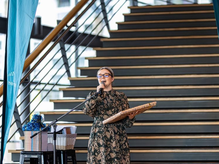 Image of A woman in a patterned dress speaks into a microphone whilst holding a wrapped present. She stands in front of a staircase, with gift hampers and blue decorations beside her.