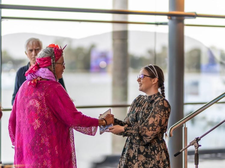 Image of A woman in a bright pink outfit and headscarf shakes hands with a smiling woman in a floral dress, as she receives an award indoors near large windows. Two other people stand in the background.
