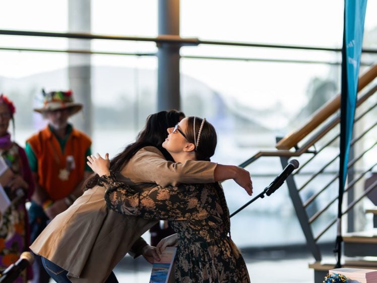 Image of Two women embrace on stage during an indoor event, whilst a few people in colourful clothing stand in the background. A microphone and staircase are visible nearby. Natural light streams through large windows.
