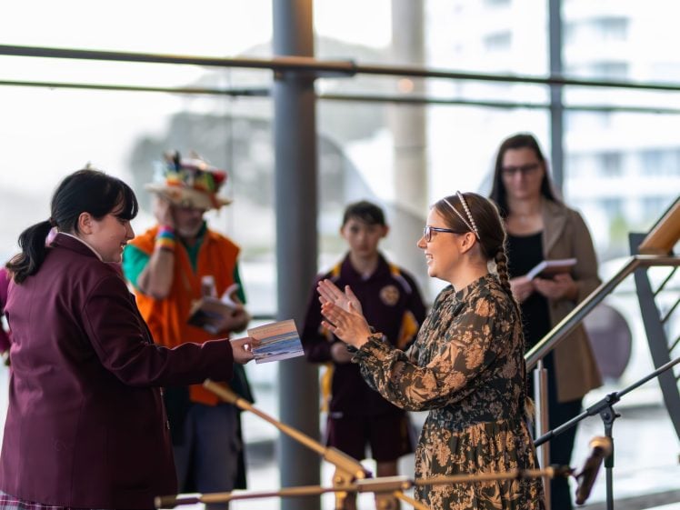 Image of A woman in patterned dress claps and smiles as she receives a stack of books from a girl in a maroon school uniform. Others, including a person in a colourful hat, watch in the background inside a bright, modern space.