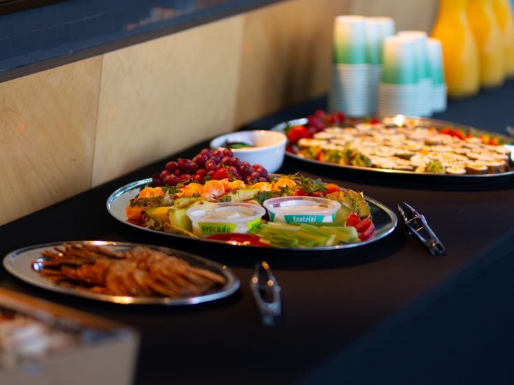 Image of A table with platters of assorted appetisers, including grapes, vegetables with dips, sliced cheese, and biscuits. Behind the food are stacked cups and jugs of orange juice.