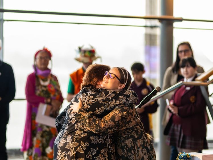 Image of Two women warmly hug each other at an indoor event, with several people standing in the background. The atmosphere appears joyful and supportive, and a microphone is visible in the foreground.