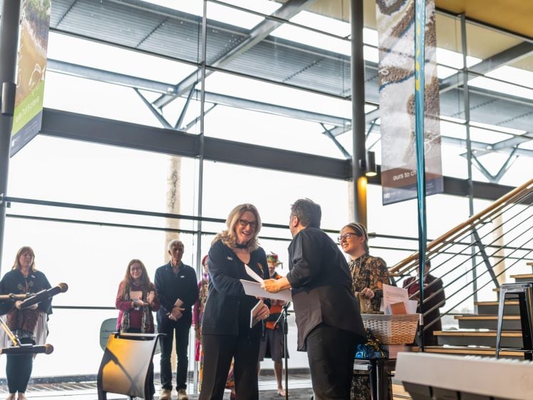 Image of A woman smiles as she receives a certificate from another person in a bright, modern indoor space with large windows. Several people stand and watch in the background near a staircase.