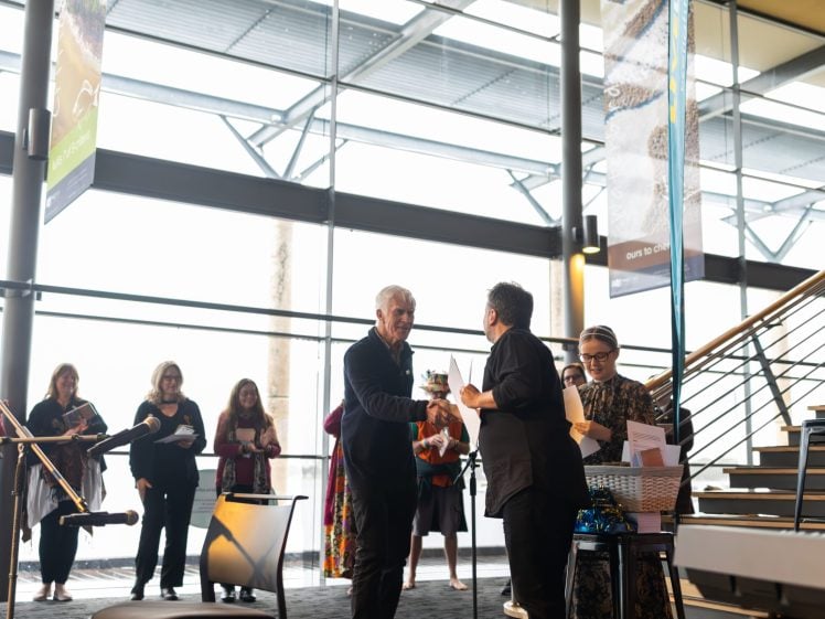 Image of Two people stand and shake hands in a bright, modern indoor space while others look on. A person holds a microphone and another holds a basket of papers nearby. Large windows and banners are visible in the background.