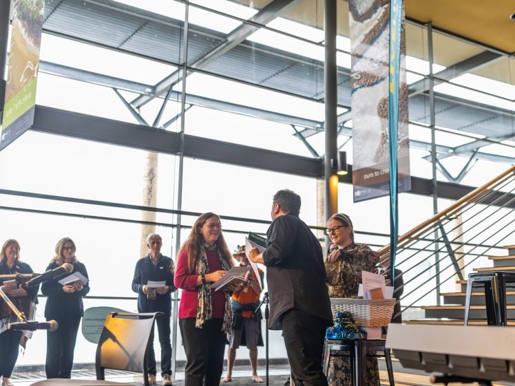 Image of A group of people stand and talk in a modern, sunlit building near a staircase. Two people in the centre exchange papers, while others observe and hold documents. Banners and large windows are visible in the background.