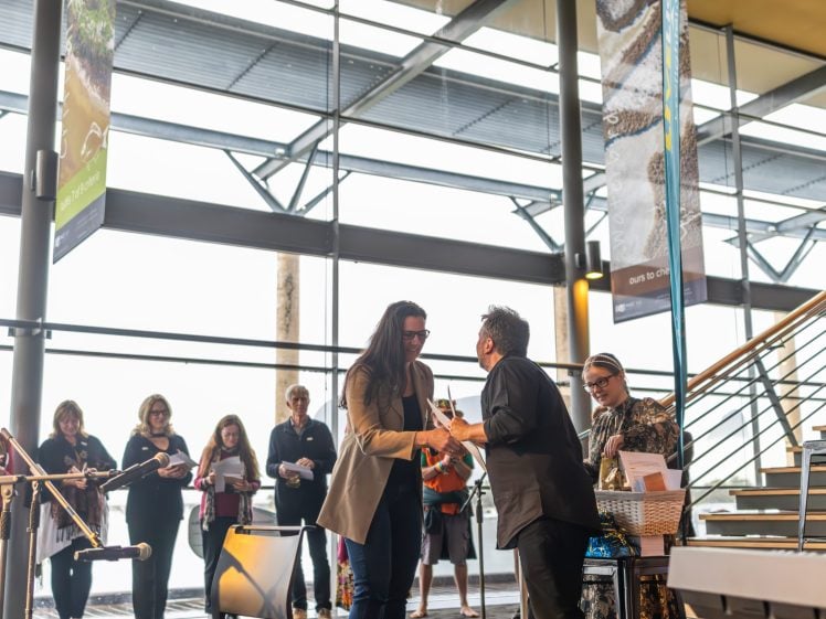 Image of Two women shake hands and smile during an indoor event, with people standing and observing in the background. A person stands at a lectern nearby. The setting is modern with large windows and banners hanging.
