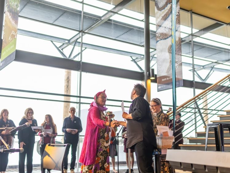 Image of A group of people gather indoors near large windows; one person in bright pink traditional attire shakes hands with another, whilst others look on and smile by a staircase.