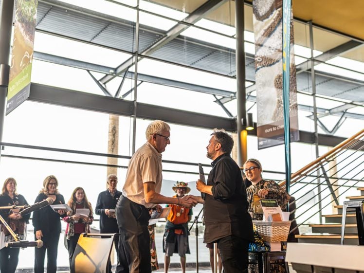 Image of Two men shake hands on stage at an indoor event, whilst a group of people watch and smile in the background. Large windows and banners are visible, allowing sunlight to fill the modern space.
