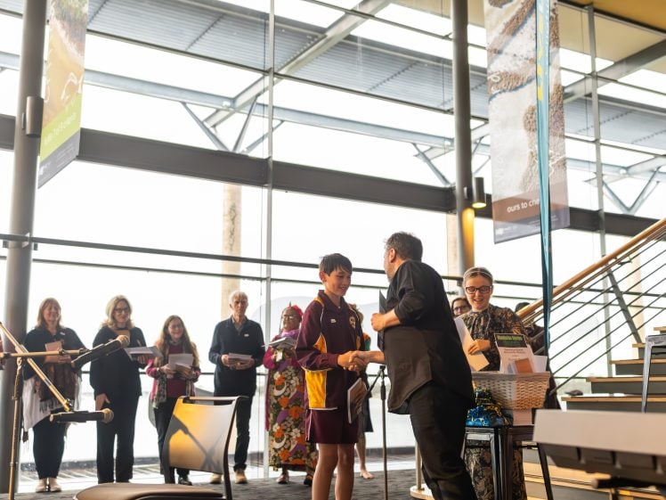 Image of A student in a maroon and yellow uniform shakes hands with an adult at an indoor awards ceremony, whilst several people stand in a queue holding certificates in the background.