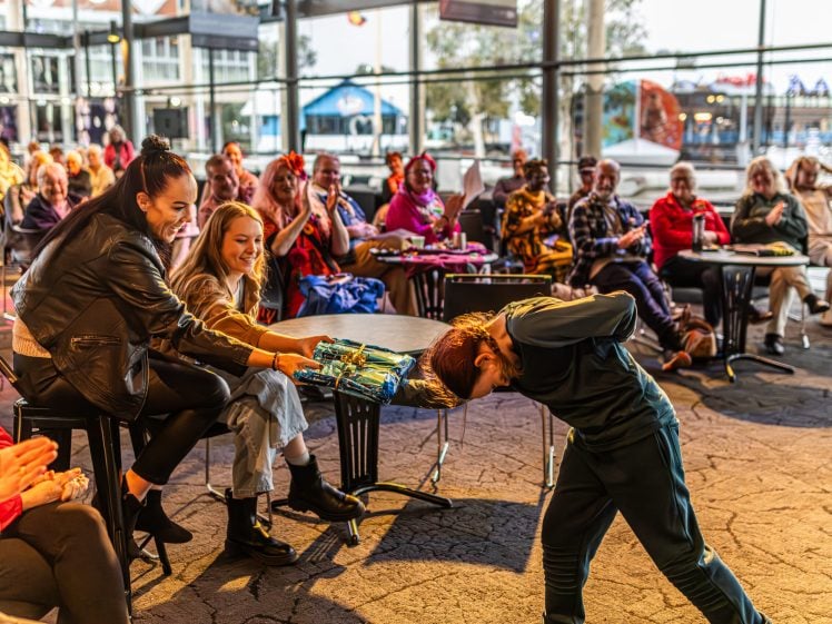 Image of A smiling woman hands a gift bag to a young boy bowing forward, whilst a seated audience in a bright, modern venue applauds and watches the interaction.
