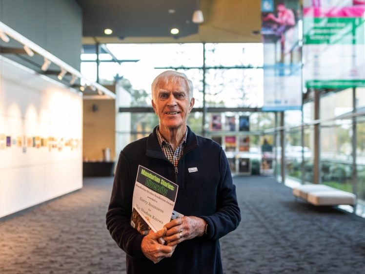 Image of An older man stands indoors, smiling and holding a book titled “Understanding Charitable Organisations: Utopia or Dystopia?” Large windows and display boards are visible in the brightly lit background.