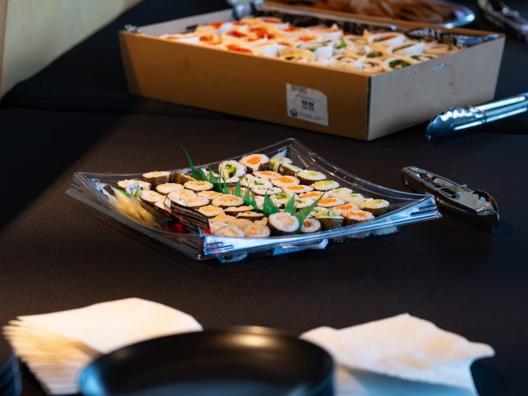 Image of A clear plastic tray filled with assorted sushi rolls is placed on a black tablecloth, with a box of more sushi in the background, metal tongs, serviettes, and black bowls nearby.