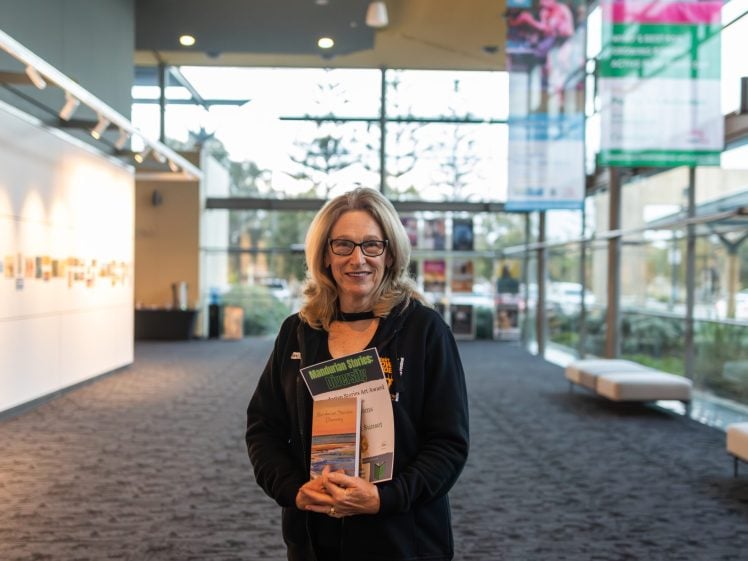 Image of A woman with blonde hair and glasses stands indoors, smiling and holding several books or brochures. She is in a modern, well-lit corridor with large windows and art displayed on the wall behind her.