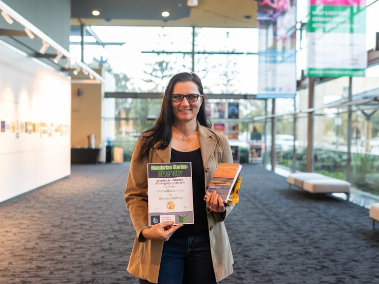 Image of A woman with long dark hair, wearing glasses and a tan blazer, stands indoors holding an award certificate and a small stack of books, smiling at the camera in a bright, modern space with large windows.