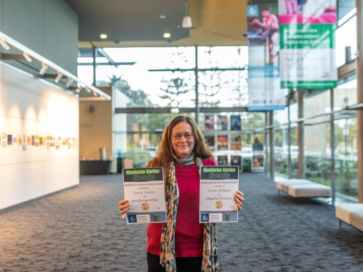 Image of A woman with long brown hair, glasses, and a colourful scarf stands indoors, smiling and holding two certificates in a brightly lit modern gallery space with large windows and posters in the background.