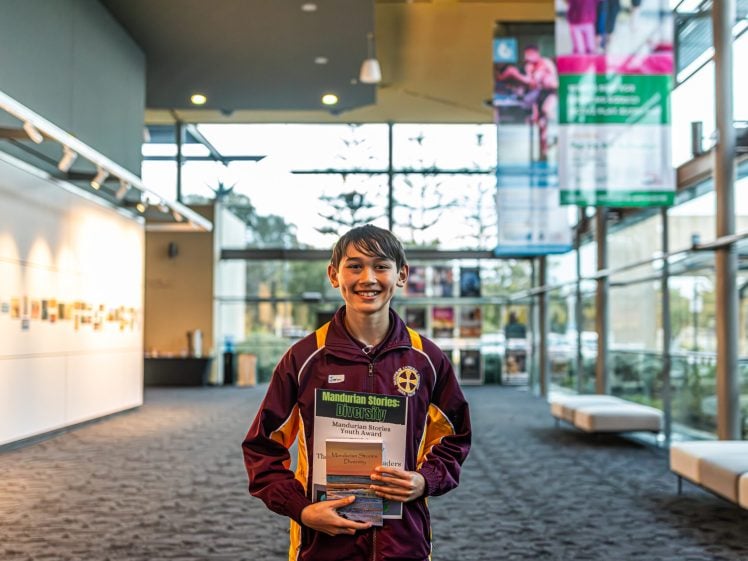 Image of A smiling student in a school jacket holding books stands in a brightly lit corridor with large windows, banners, and seating along the side.