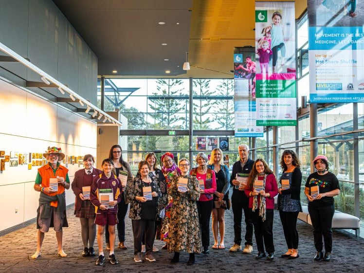 Image of A diverse group of people, some in fancy dress and school uniforms, stand indoors in a bright gallery space holding certificates and smiling at the camera. Colourful banners and photos are displayed on the walls.