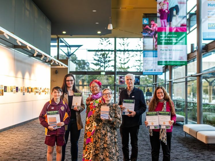 Image of Six people stand indoors, smiling and holding certificates. They are positioned in a well-lit corridor with large windows, banners, and photos displayed on the wall behind them.