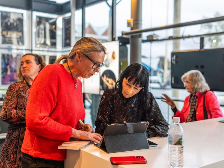 Image of Four women are gathered in a modern, well-lit indoor space. Two women in the foreground are focused on a tablet and notebook, while two others sit and talk in the background near water bottles. Posters decorate the walls.