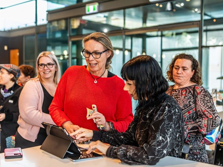 Image of A group of women stand around a table, smiling and interacting with a tablet device in a modern, glass-walled room. One woman in red holds a coffee cup, whilst another gestures towards the tablet.