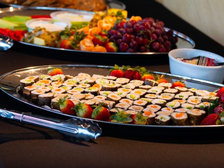 Image of A large silver platter filled with assorted sushi rolls, garnished with fresh strawberries, sits on a black tablecloth. In the background, there is another platter with fruit and vegetables, and a small bowl with condiment sachets.