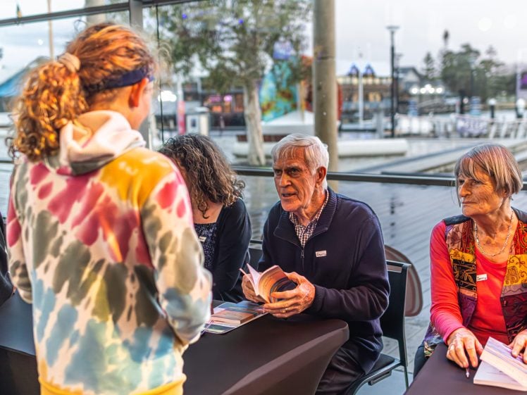 Image of A young person in a colourful hoodie talks to two older adults sitting at a table with books, possibly at a book signing event in a bright, modern indoor space with large windows overlooking a waterfront.