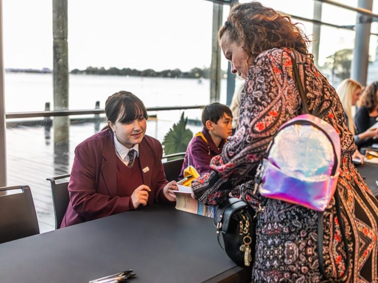 Image of A student in a maroon uniform sits at a table, signing a book for a woman with a colourful bag. Another student sits nearby. The setting is indoors with large windows overlooking water.