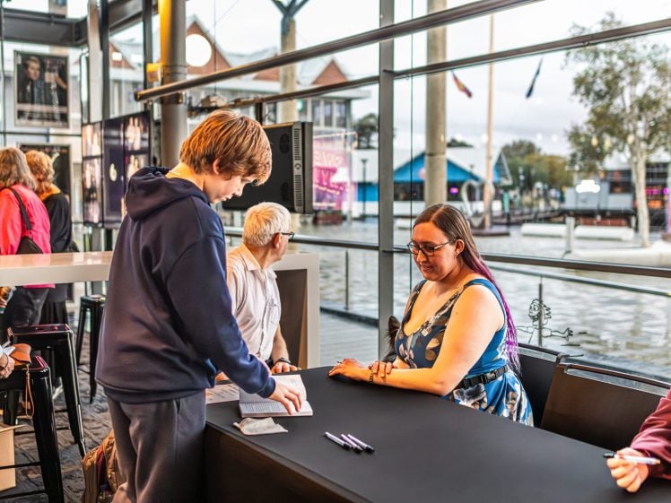 Image of A young person in a hoodie gets a book signed by a woman in a colourful dress at a table inside a bright, modern building with large windows overlooking a riverside scene. Other people wait nearby.